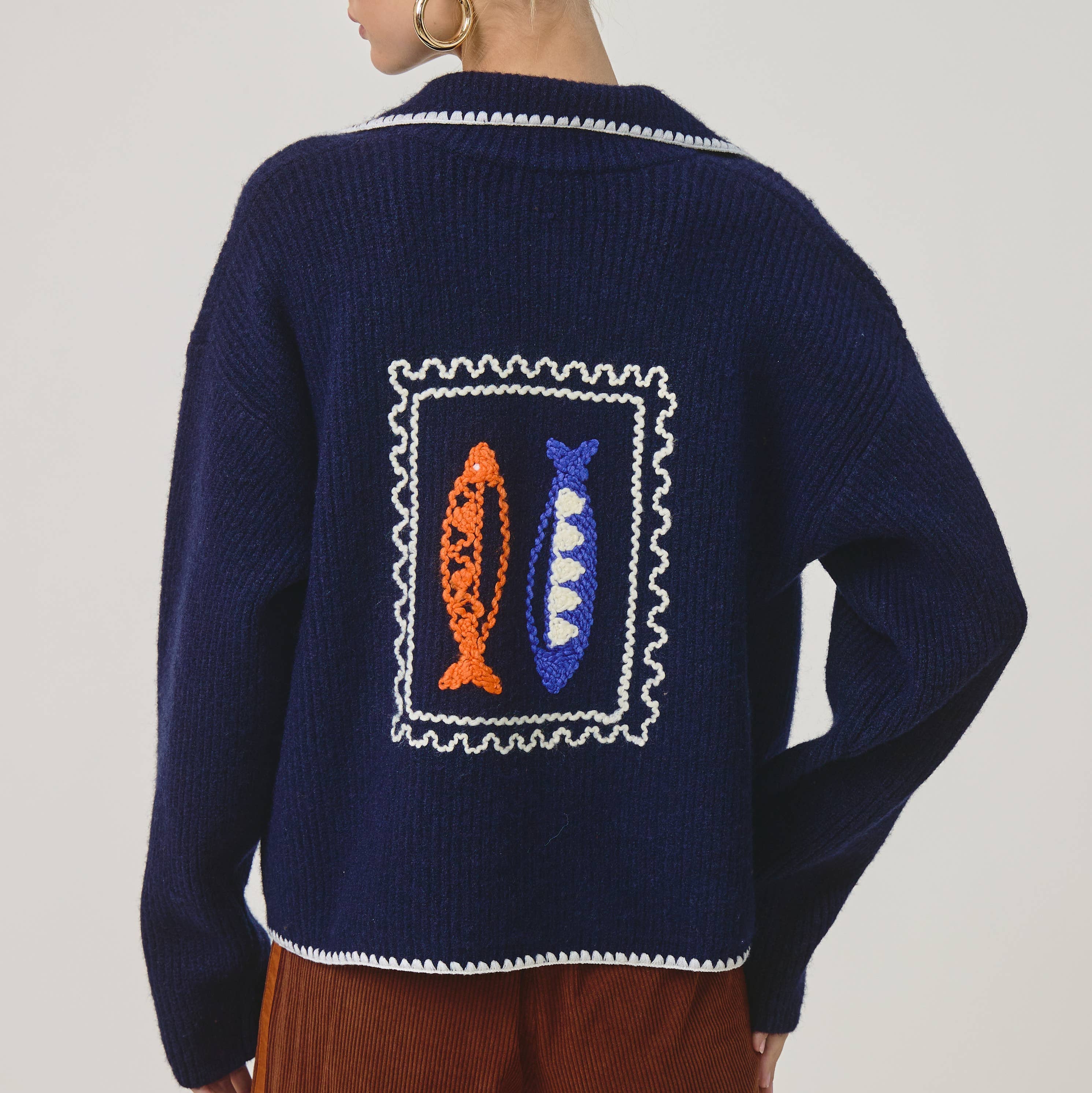 A woman shown from the back wearing a navy blue knit cardigan with a large fish patch design on the back, paired with brown pants, against a light gray studio background.