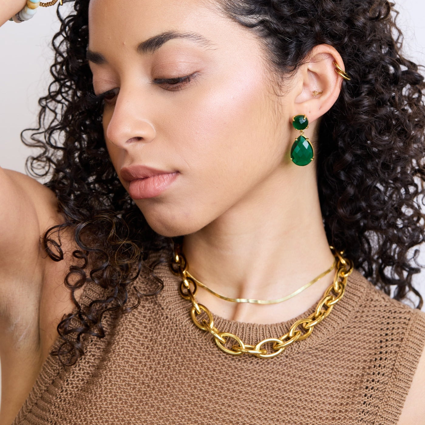 Woman with curly hair wearing green earrings and gold necklaces against a white background