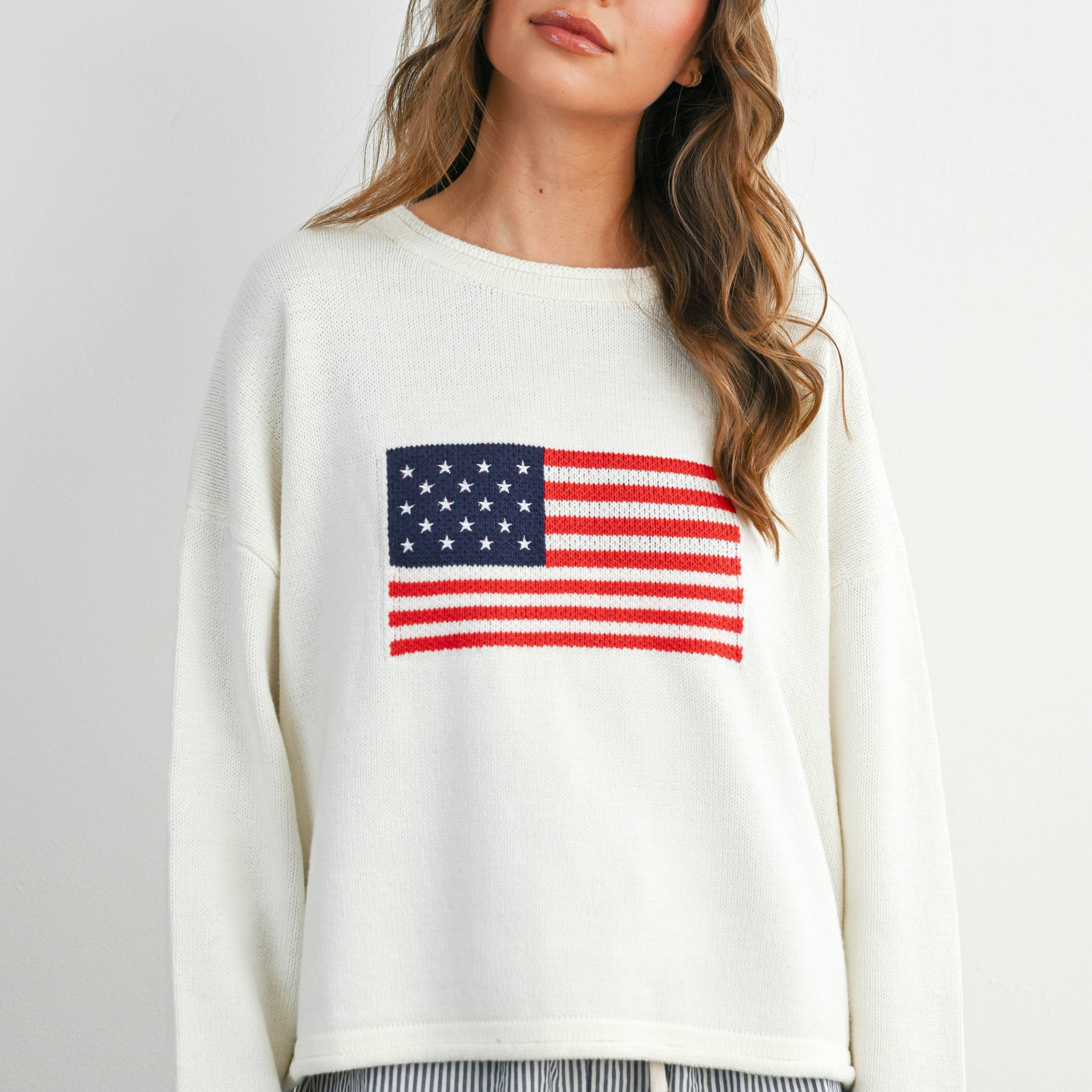 Woman wearing a white sweatshirt with an American flag design and striped shorts on a plain background