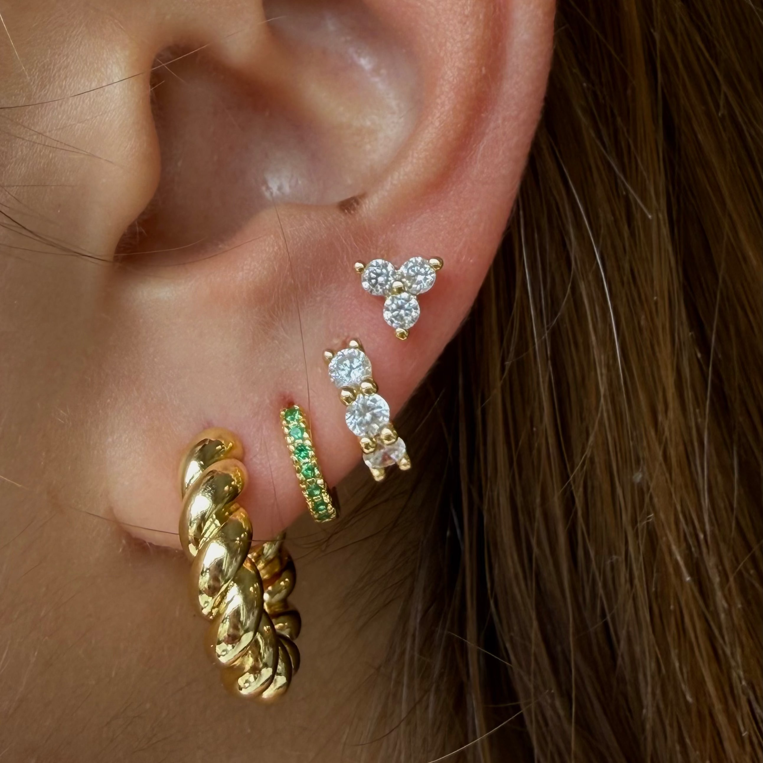 A close-up of a person's ear showcasing a stack of four gold and green stud/huggie earrings, including a twisted gold hoop, a gold huggie with green stones, and two small gold flower-shaped studs with clear crystals.